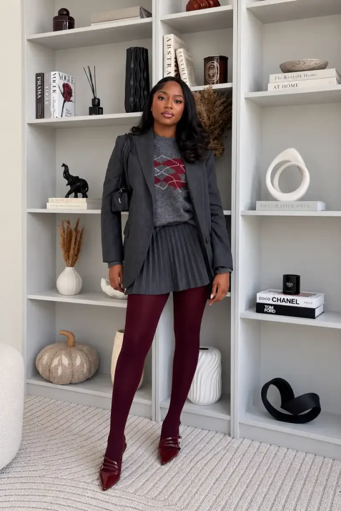 Black woman styling a charcoal blazer over a grey argyle sweater and pleated mini skirt with burgundy tights and pointed strap heels.