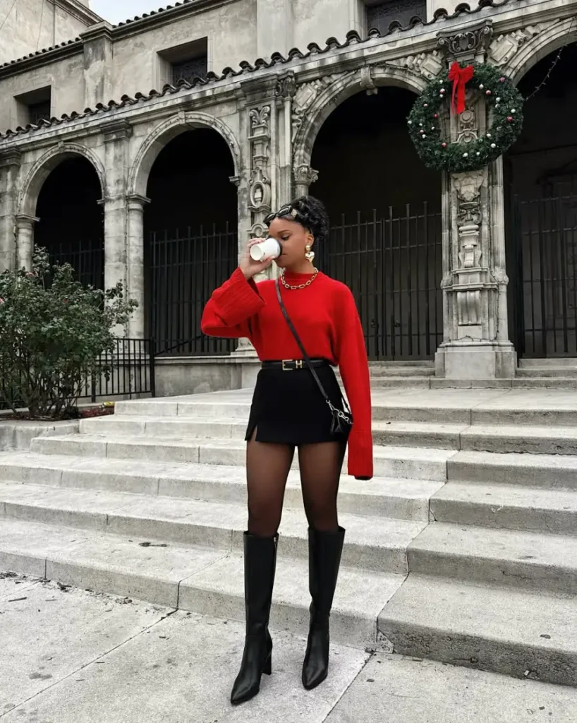 Black woman wearing a bright red sweater with a black mini skirt, sheer tights, knee-high black boots, and a small crossbody bag for a bold fall look.