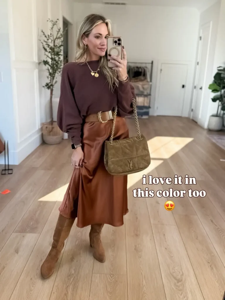 Woman wearing a plum knit sweater with a cinnamon brown satin midi skirt, tan belt, suede knee-high boots, and an olive green quilted handbag.