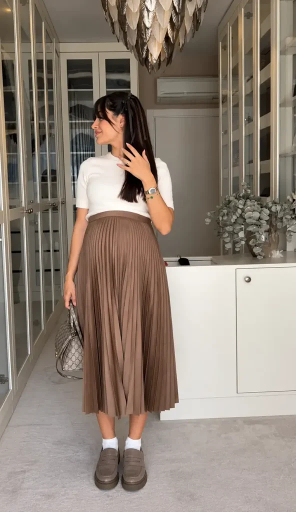 Pregnant woman styling a white short-sleeve knit top with a taupe pleated midi skirt, brown loafers, and a patterned handbag for a cute maternity fall outfit.