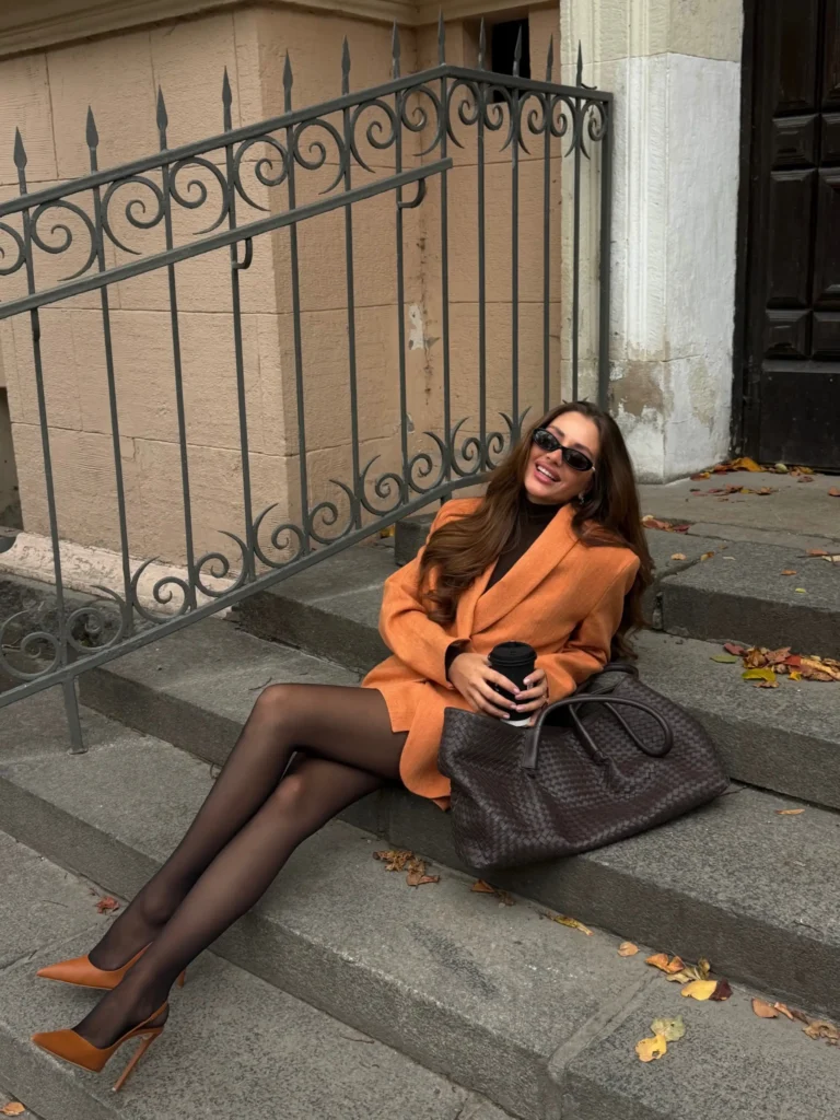 Woman wearing a warm orange blazer-style mini outfit over a black turtleneck with sheer tights, brown pointed heels, and a dark woven bag for fall.