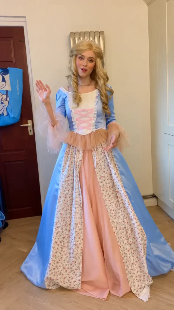 Woman wearing a blue and blush storybook princess gown with floral skirt panels, pink corset details, sheer sleeves, curls, and a tiara.