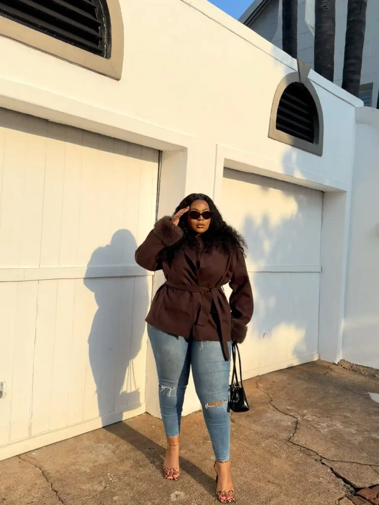 Curvy Black woman wearing a belted chocolate brown jacket with furry cuffs, distressed blue skinny jeans, leopard-print heels, and a black handbag for a cute casual fall look.