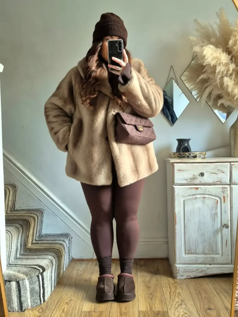Plus size woman styling an oversized taupe faux fur coat with chocolate brown leggings, a knitted beanie, brown ankle boots, and a quilted crossbody bag.