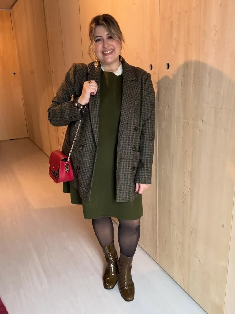 Plus size woman wearing an olive green dress with a checked blazer, black tights, brown ankle boots, and a red handbag for fall.