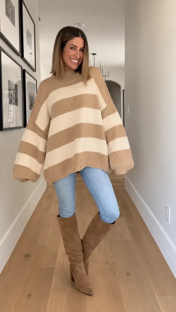 Woman wearing an oversized camel and cream striped sweater with light blue skinny jeans and tall tan suede boots for a cozy Thanksgiving outfit with jeans.