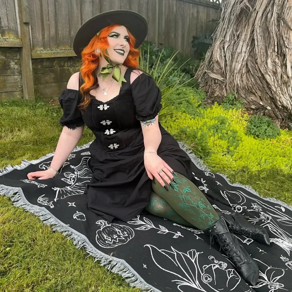Plus size woman in a black woodland witch dress with puff sleeves, wide-brim hat, green bow, and patterned tights.
