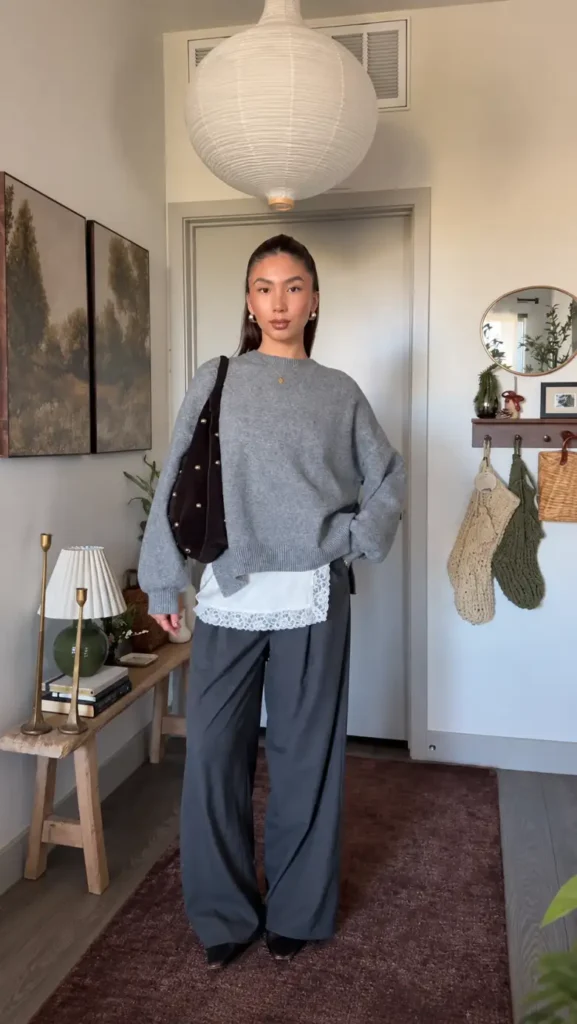 Woman wearing a grey oversized sweater over a white lace-trim layer with dark wide-leg trousers and a brown shoulder bag.