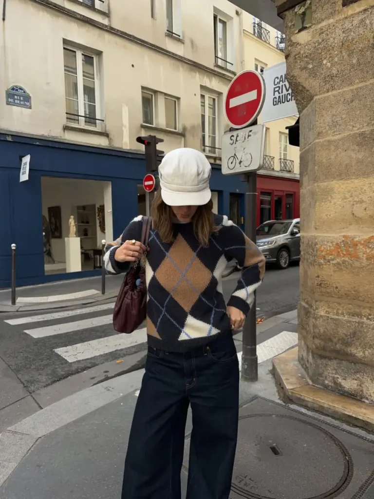 Woman wearing a navy and tan argyle sweater with dark wide-leg jeans, a cream cap, and burgundy shoulder bag for a casual autumn street style look.