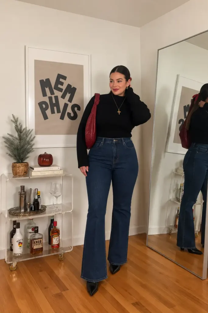 Woman wearing a fitted black turtleneck with dark-wash flared jeans, black pointed-toe boots, gold jewelry, and a burgundy shoulder bag for a classy Thanksgiving dinner outfit.