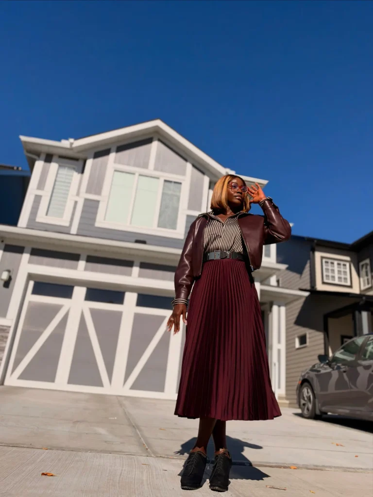 Black woman styling a burgundy pleated midi skirt with a striped blouse, cropped leather-look jacket, wide belt, and chunky black sneakers for a casual fall look.