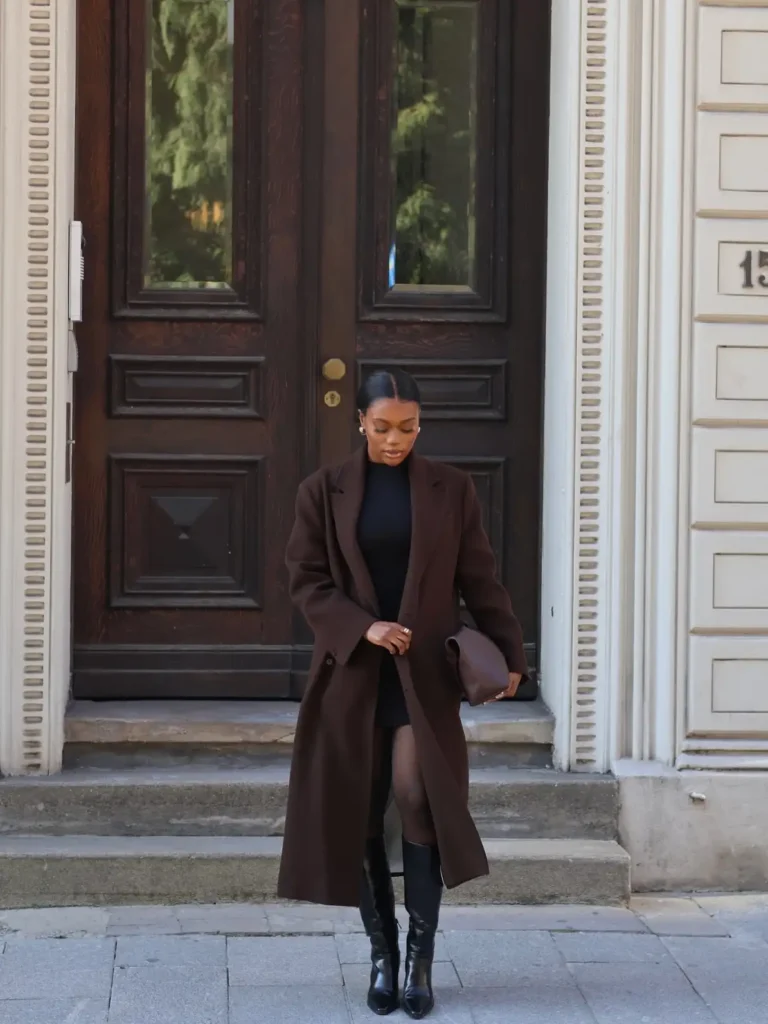 Black woman wearing a long espresso brown coat over a short black dress with sheer tights, knee-high black boots, and a brown clutch for an elegant fall outfit.