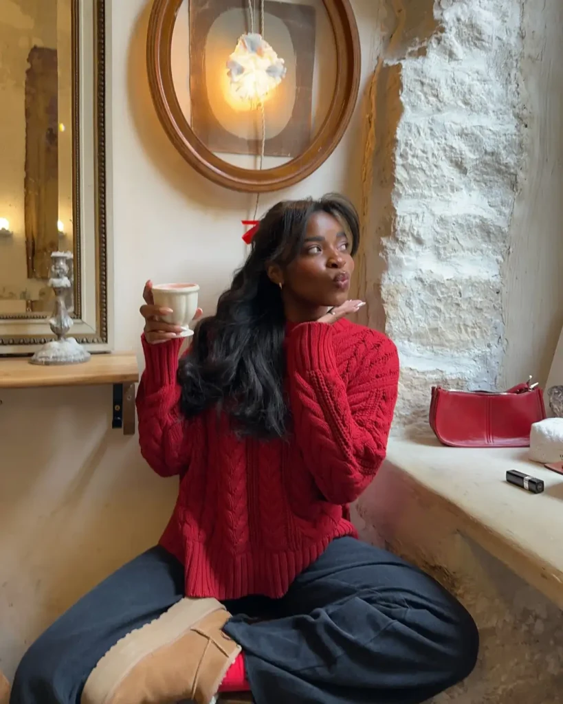 Black woman wearing a chunky cherry red cable-knit sweater with relaxed charcoal gray trousers, tan ankle boots, red socks, and a matching red hair bow for a cozy winter look.