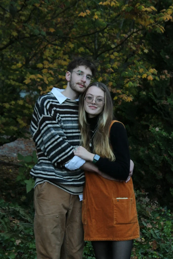Couple posing in autumn outfits with a rust corduroy pinafore dress, black turtleneck, striped oversized sweater, and relaxed brown trousers.