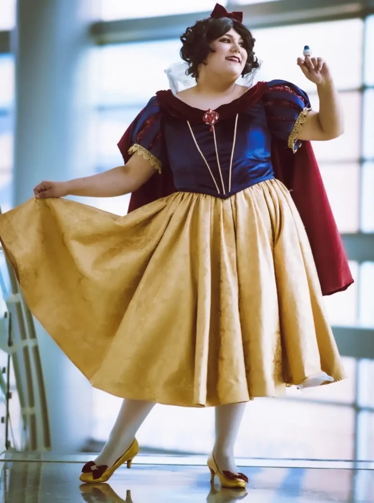 Plus size woman wearing a fairytale princess Halloween costume with a blue bodice, golden skirt, red cape, and bow.