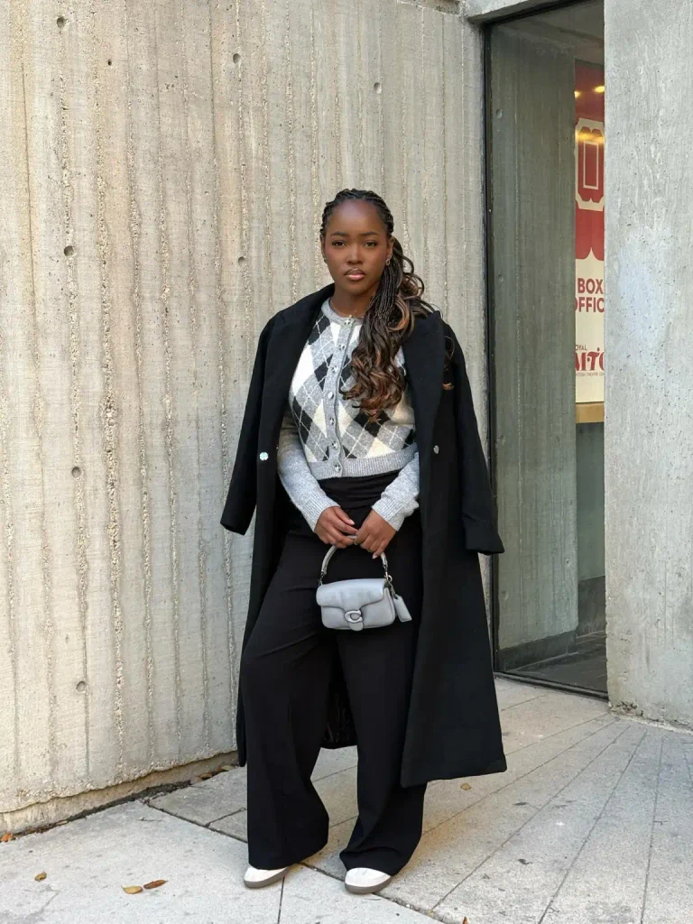 Black woman wearing a gray argyle cardigan with black wide-leg trousers, a long black coat, white sneakers, and a small gray handbag for casual office style.