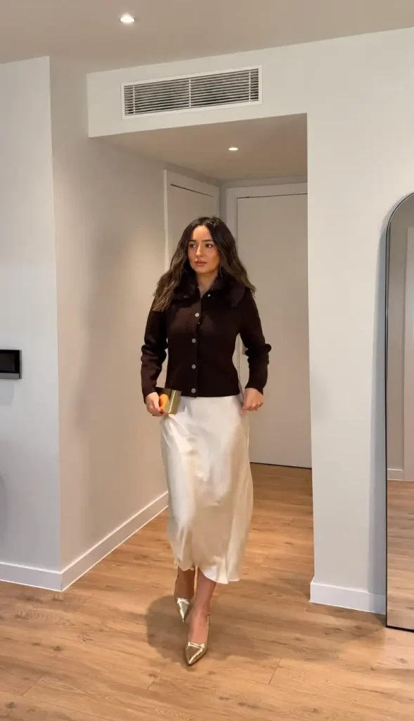Woman styling an ivory satin midi skirt with a dark chocolate cardigan featuring a plush collar, metallic gold pointed heels, and a gold clutch for an elegant winter dinner look.