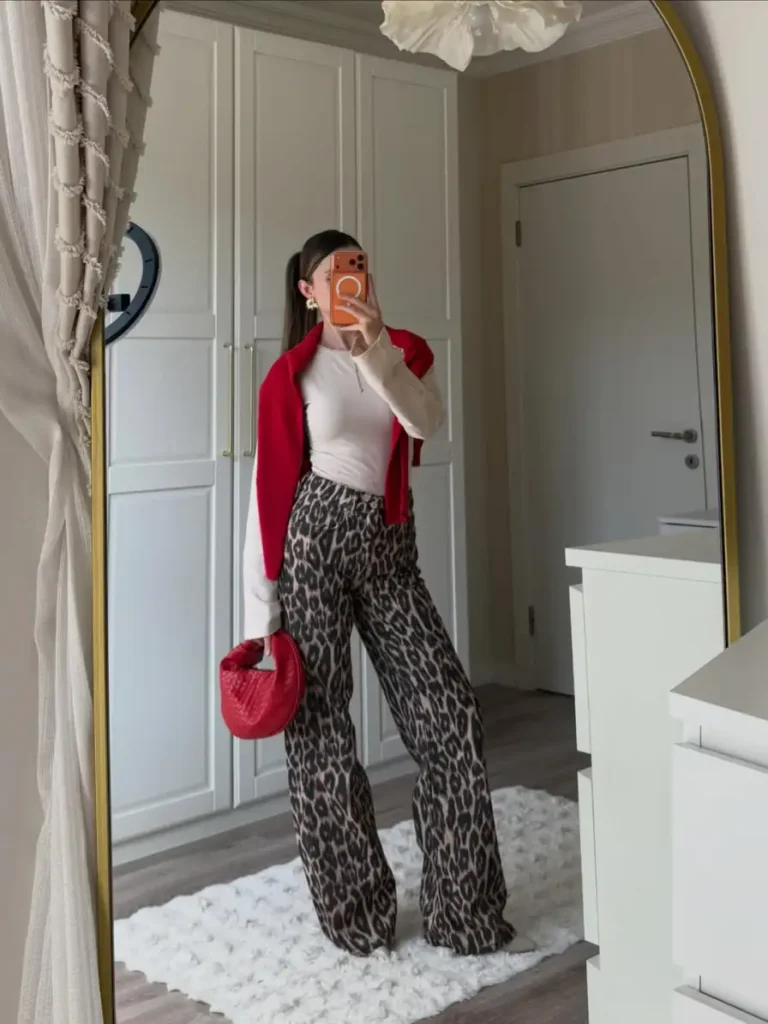 Woman wearing wide-leg leopard print trousers with a fitted cream top, red sweater over the shoulders, and a matching red handbag for a stylish fall outfit.