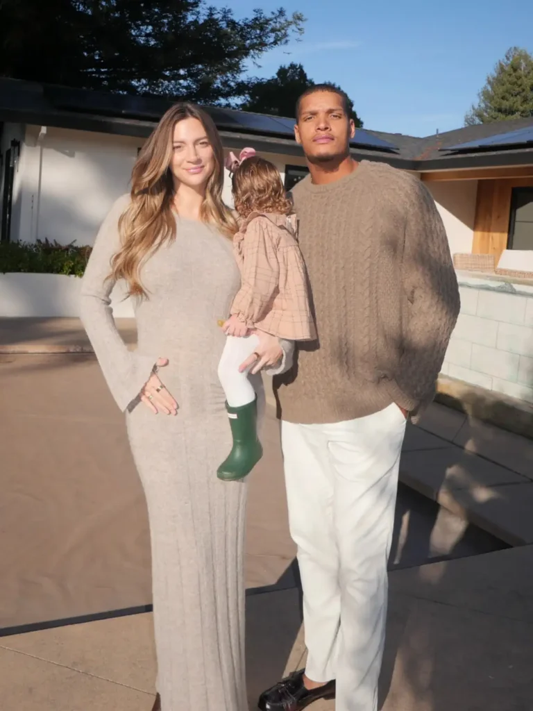 Mom wearing a gray knit maxi dress, Dad in a taupe cable-knit sweater and cream trousers, and their daughter in a dusty pink checked outfit with green rain boots.