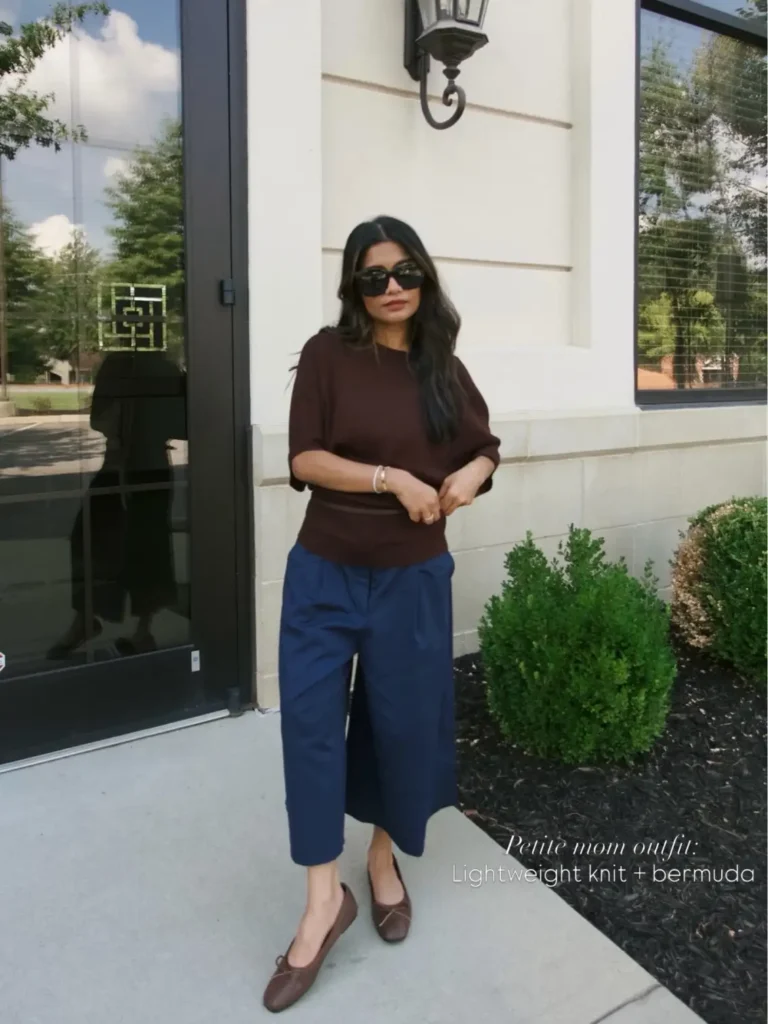 Petite woman wearing a brown lightweight knit top with navy Bermuda shorts, brown ballet flats, and dark sunglasses for an early fall outfit.