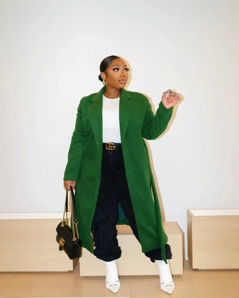 Plus size Black woman wearing a long green coat, white top, black cargo pants, and pointed white boots for a modern casual fall look.