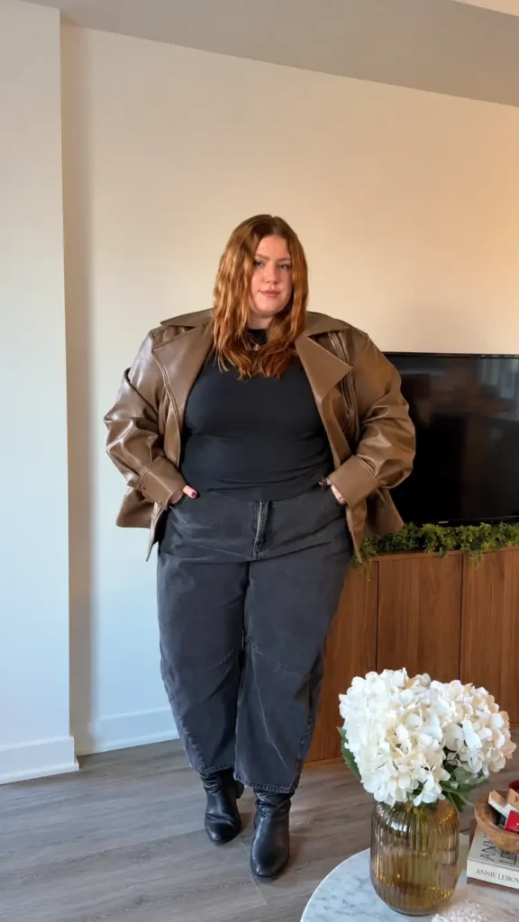 Plus size woman wearing an oversized brown leather jacket with a black top, charcoal wide-leg jeans, and black ankle boots for an edgy winter street-style outfit.