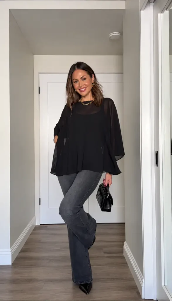 Woman wearing a flowing sheer black blouse with wide sleeves, charcoal gray jeans, pointed black heels, and a quilted handbag for a dressy casual Thanksgiving look.