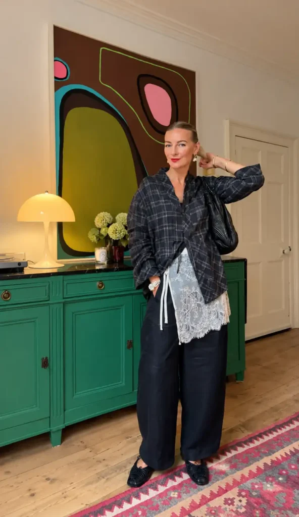 Stylish mature woman wearing an oversized dark plaid shirt layered over a white lace top with loose black trousers and woven black shoes for a trendy Thanksgiving outfit.