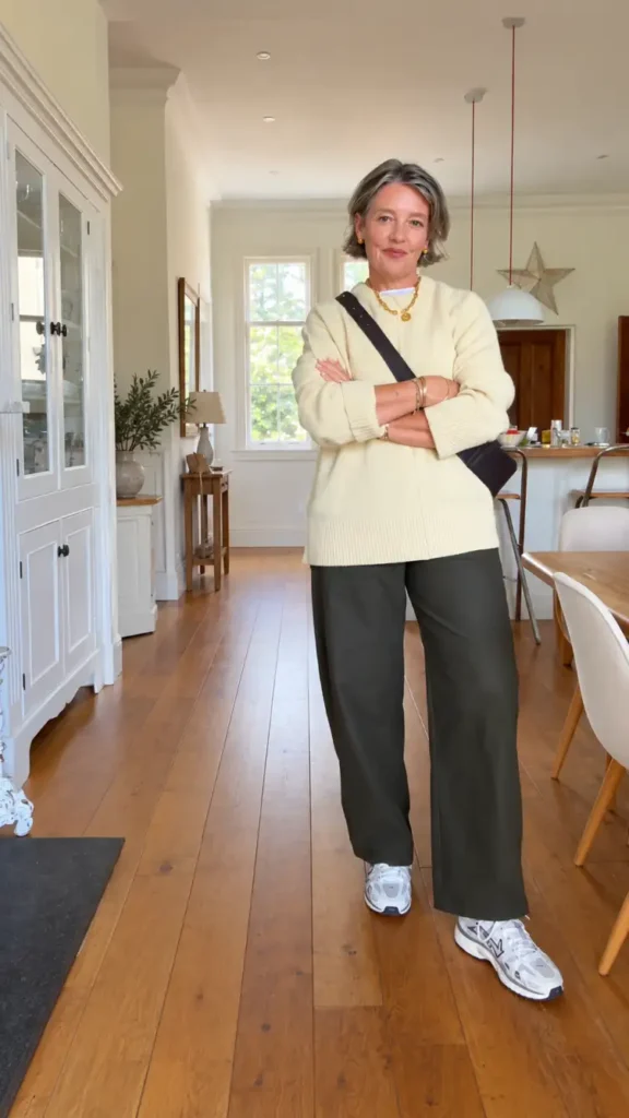 Woman over 50 wearing a butter-yellow oversized sweater, dark olive wide-leg trousers, white and gray sneakers, and a crossbody bag for a casual winter look.