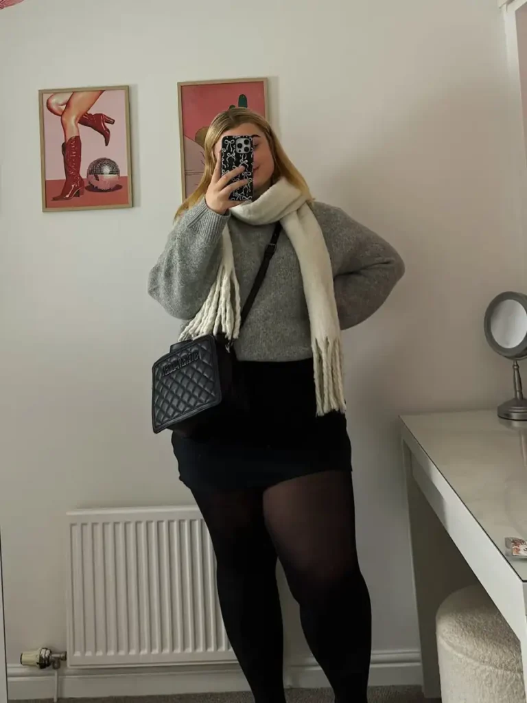 Plus size fall outfit with an oversized grey knit sweater, black mini skirt, sheer black tights, cream scarf, and quilted black crossbody bag.