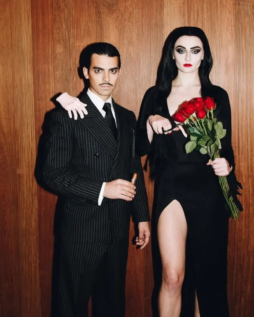 Spooky couple Halloween costumes with a black pinstripe suit, long black dress, pale gothic makeup, and red roses.