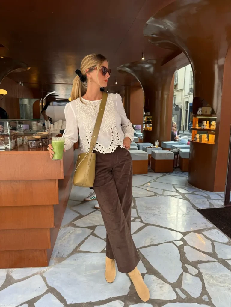 Woman wearing a white eyelet lace blouse with brown wide-leg trousers, tan ankle boots, sunglasses, and a crossbody bag.