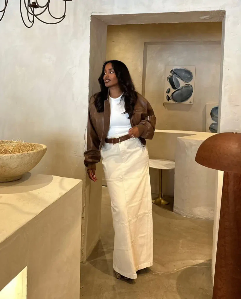  Black woman styling a brown leather bomber jacket over a white T-shirt and ivory maxi skirt with a brown belt for a casual autumn street style look.