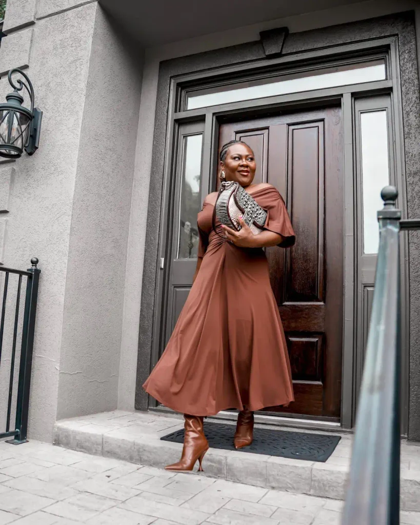 Black woman wearing a chocolate-brown off-shoulder midi dress with gathered details, cognac knee-high boots, and a patterned clutch for an elegant autumn outfit.