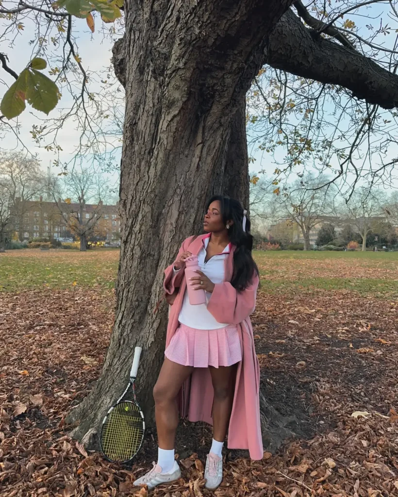 Black woman wearing a long pink coat over a white quarter-zip top and pink pleated tennis skirt, paired with white socks, pastel sneakers, and a pink tumbler.