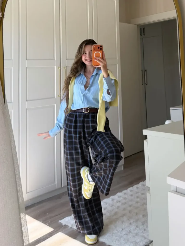 Woman wearing a light blue button-down shirt with a yellow sweater over her shoulders, plaid wide-leg trousers, and yellow sneakers for a playful fall outfit.