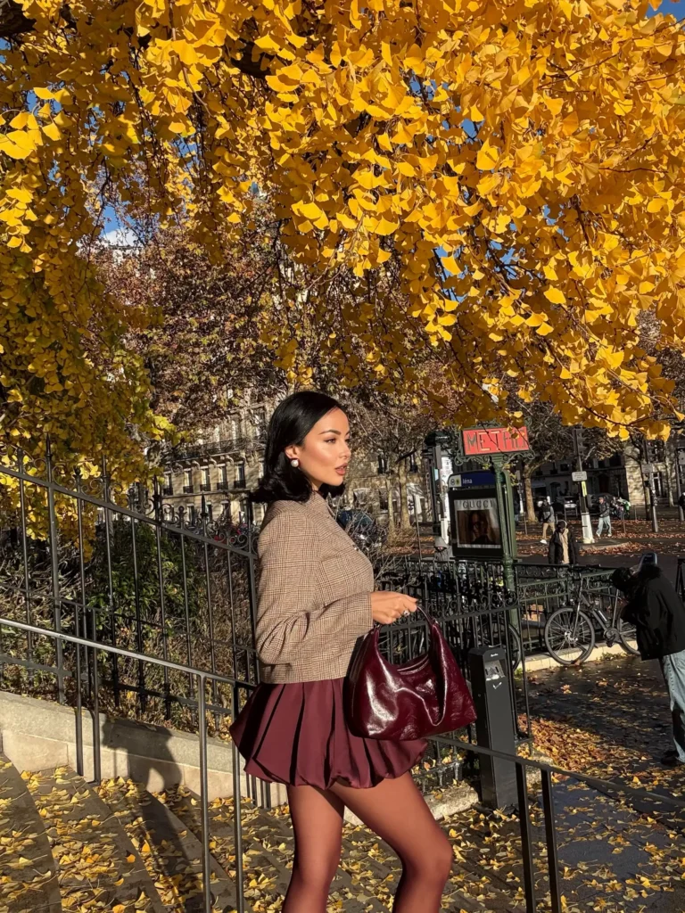 Woman wearing a checked fitted top with a burgundy pleated mini skirt and glossy burgundy handbag for a feminine autumn city look.