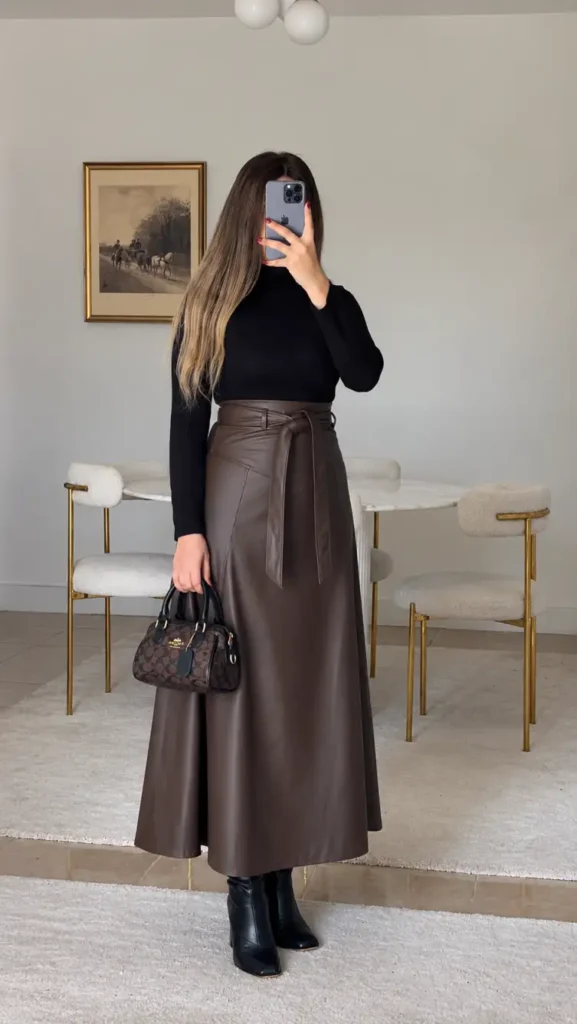 Woman wearing a chocolate-brown leather maxi skirt with a belted waist, fitted black turtleneck, black ankle boots, and a small patterned handbag for an elegant winter dinner outfit.