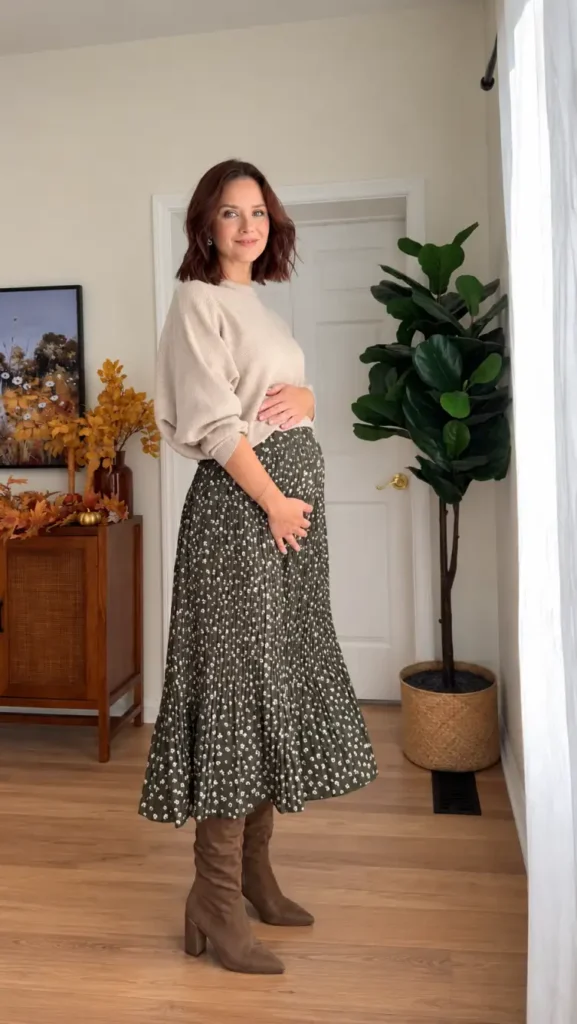 Pregnant woman wearing a beige oversized sweater with an olive green floral maxi skirt and tall brown suede boots for a cozy fall maternity outfit.