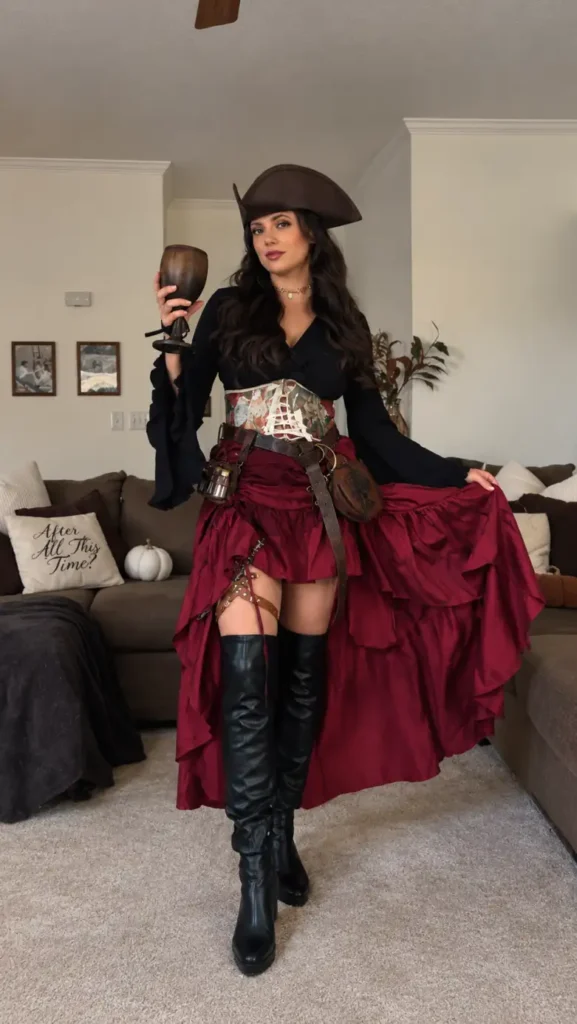 Woman wearing a black blouse, patterned corset, layered burgundy skirt, pirate hat, utility belts, and tall black boots for Halloween.