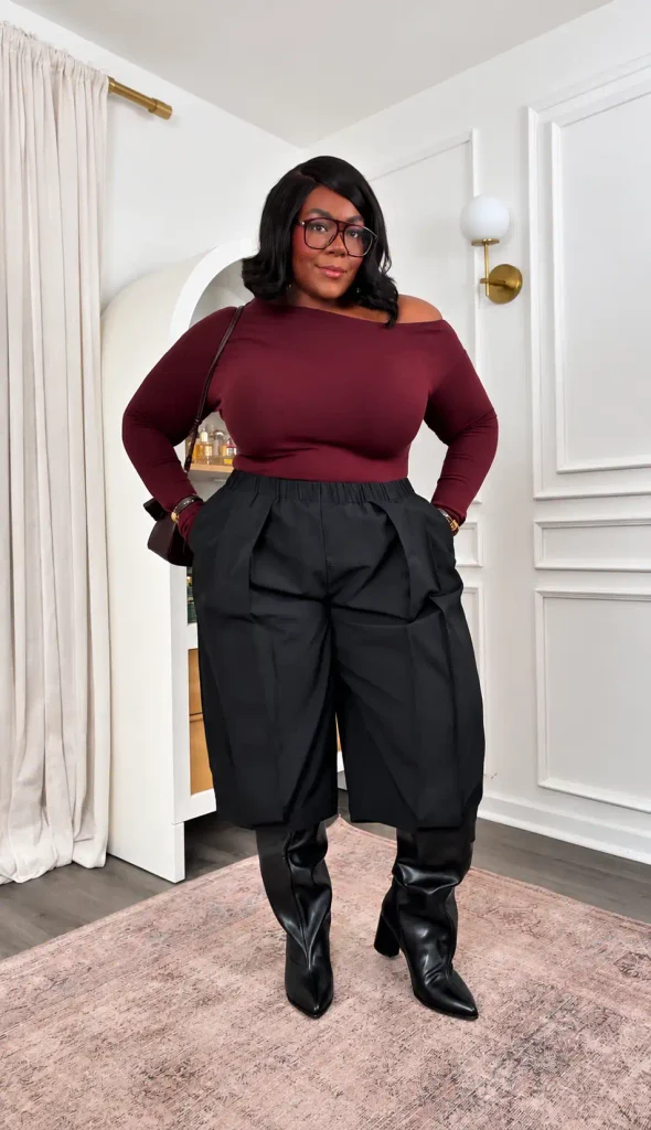 Curvy Black woman wearing a burgundy off-shoulder top with black pleated Bermuda shorts, knee-high leather boots, and a small shoulder bag for a trendy fall outfit.