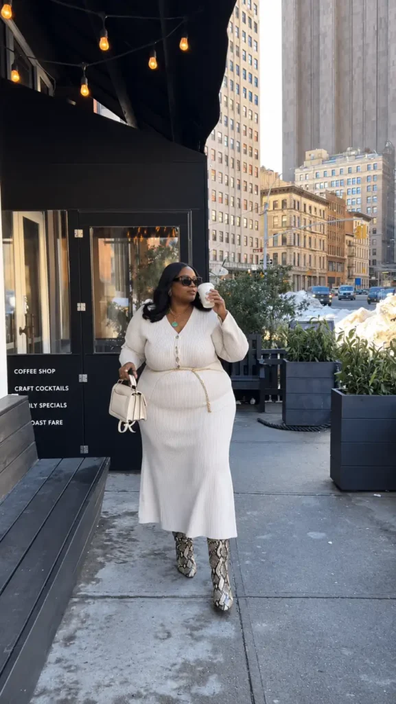 Plus size Black woman wearing a cream knit top and matching ribbed maxi skirt with a gold chain belt and snakeskin boots for fall.