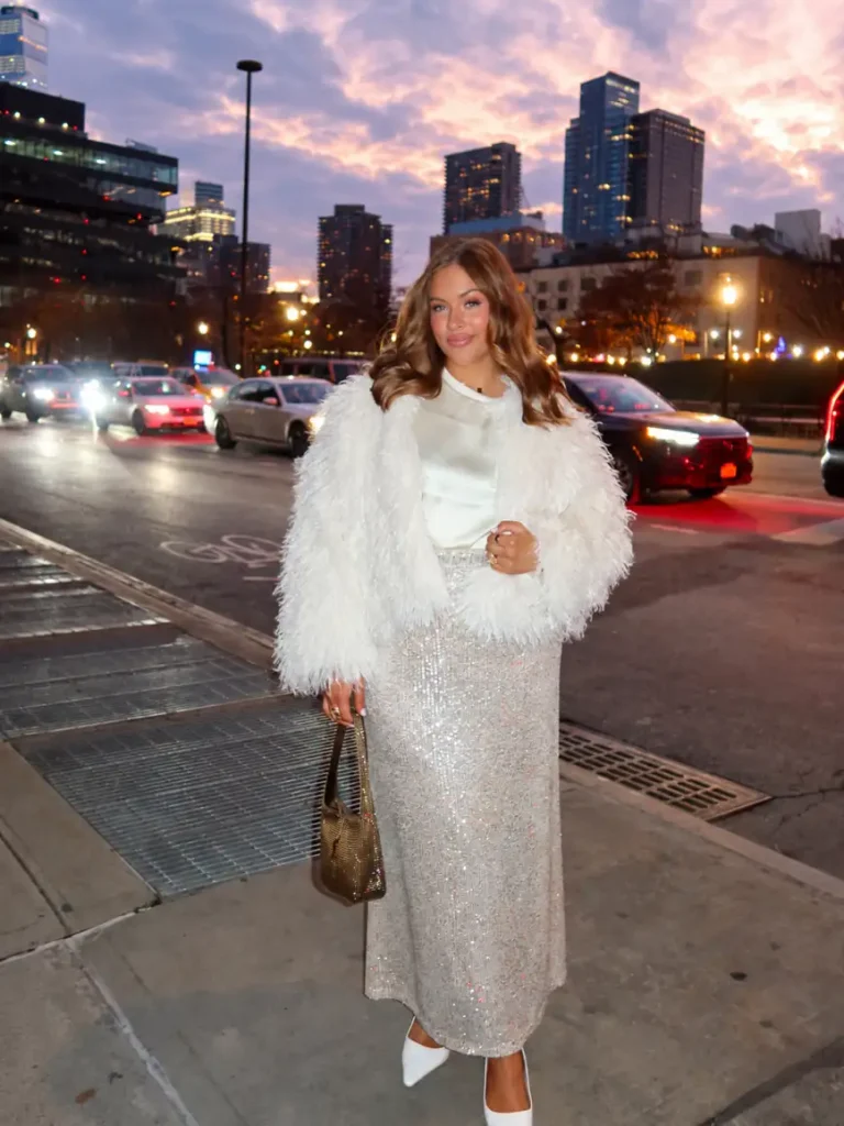 Plus-size woman styling a fluffy white cropped jacket over an ivory satin top and silver sequin midi skirt with white pointed heels for an elegant holiday outfit.