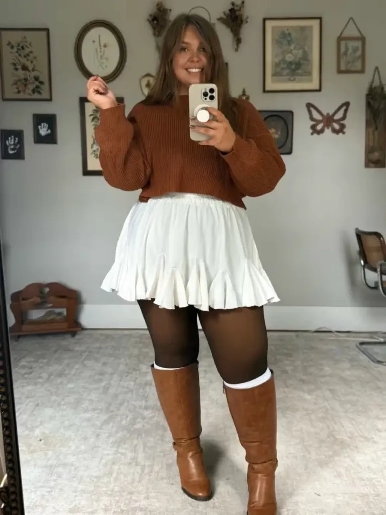 Plus size woman wearing a rust brown sweater with a white flared mini skirt, brown tights, and tall cognac boots for a cute fall outfit.