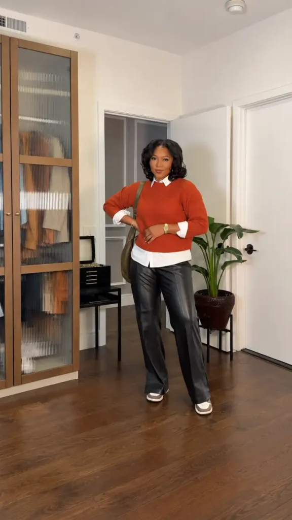 Black woman wearing a rust-orange sweater layered over a white collared shirt with black leather-look trousers and neutral sneakers for a business casual fall outfit.