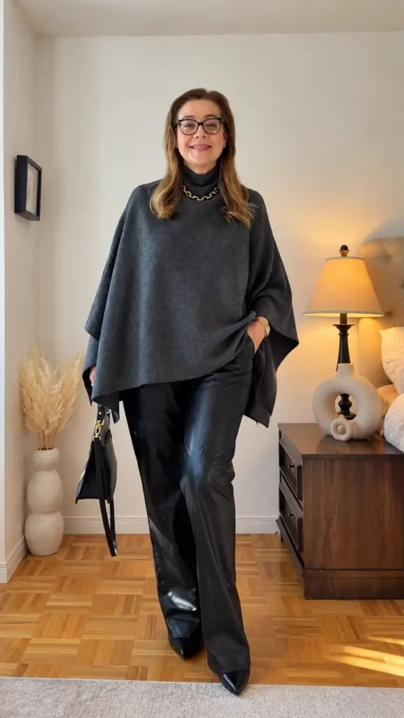 Woman over 50 wearing a charcoal gray poncho-style sweater with black leather trousers, pointed black shoes, and gold chain jewelry for an elegant Thanksgiving dinner look.