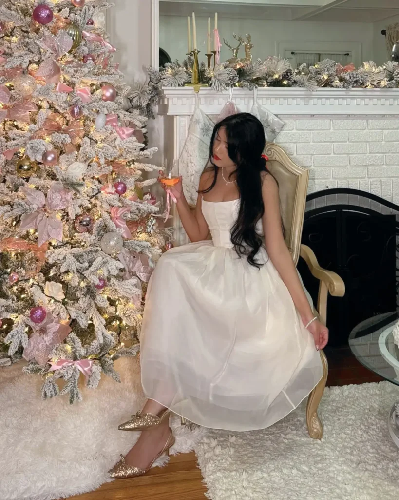 Woman wearing an ivory strapless corset midi dress with a full flowing skirt and glittering gold slingback heels beside a pink decorated Christmas tree.