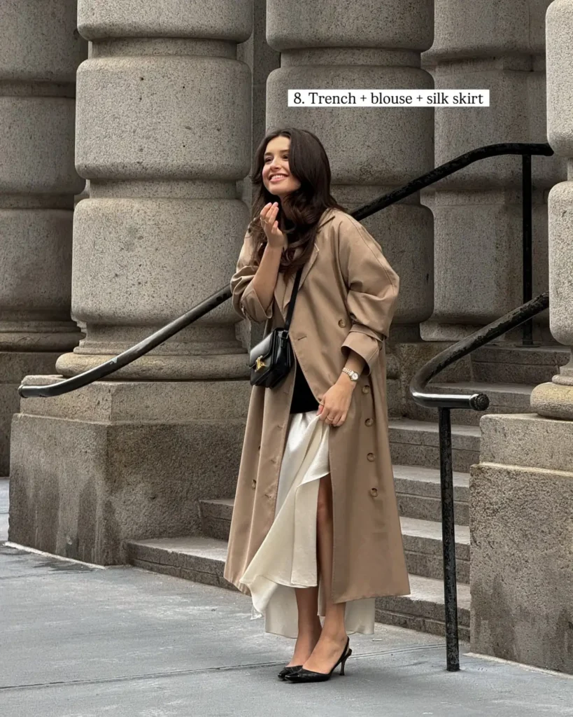Camel trench coat layered over a black top and ivory satin skirt with black slingback heels and a small black bag.