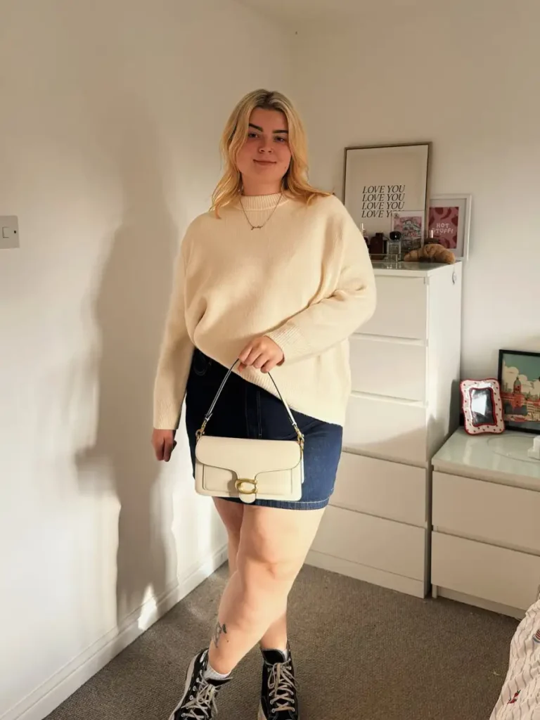 Plus size fall outfit with an oversized cream sweater, dark denim mini skirt, black high-top sneakers, and a structured cream shoulder bag.