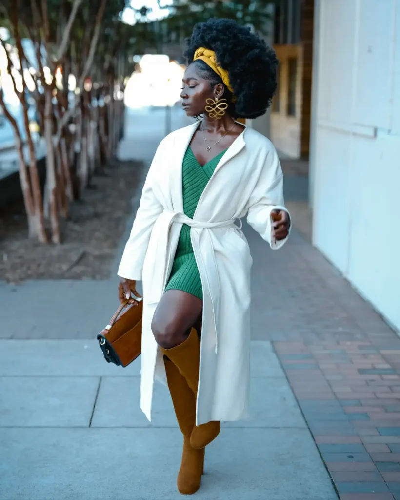Black woman wearing an emerald green knit mini dress with a long ivory wrap coat, camel over-the-knee boots, brown handbag, yellow headwrap, and statement earrings.